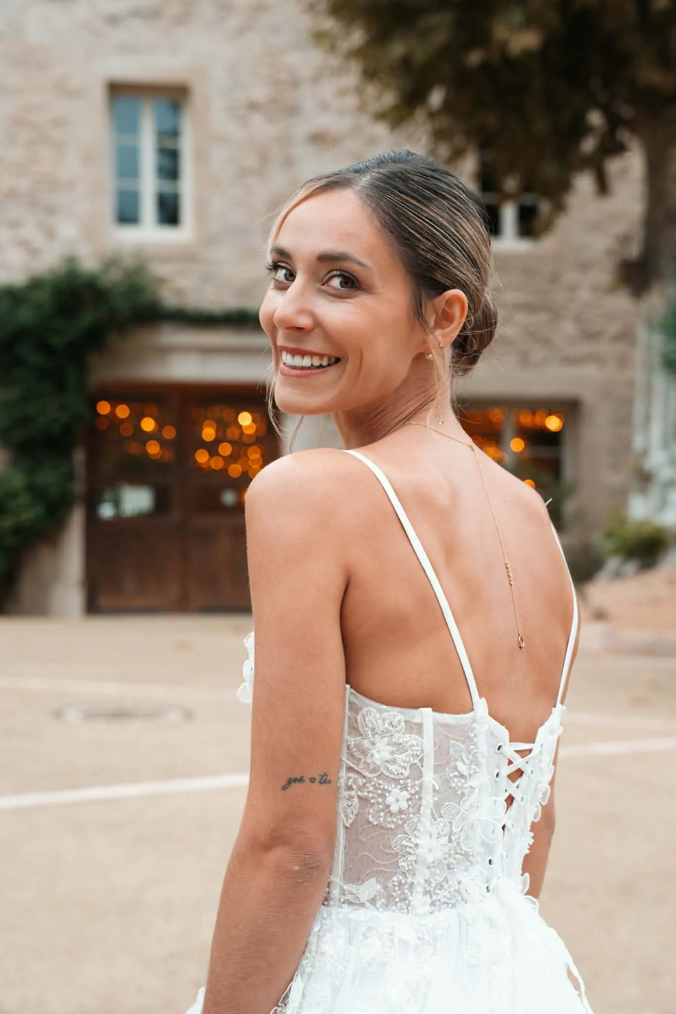 Mariée souriante de dos devant une bâtisse en pierre, chignon bas et robe en dentelle brodée